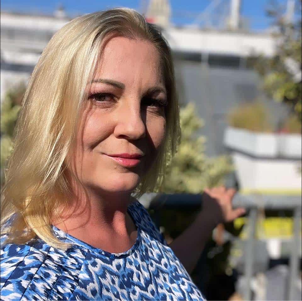 Frau mit blonden Haaren steht draußen in der Sonne und lächelt sanft. Sie trägt ein blau gemustertes Outfit. Im Hintergrund sind verschwommene Gebäudedetails und Grünpflanzen zu sehen. Professionelles W