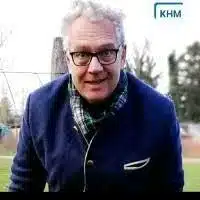 Mann mit Brille und lockigem Haar in einem blauen Sakko im Freien, mit KHM-Logo im Hintergrund, repräsentiert kreative Webdesign-Dienstleistungen in Wien.